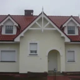 Dom jednorodzinny z nowym dachem z czerwonej dachówki ceramicznej, białymi oknami dachowymi i ozdobną konstrukcją szczytową.